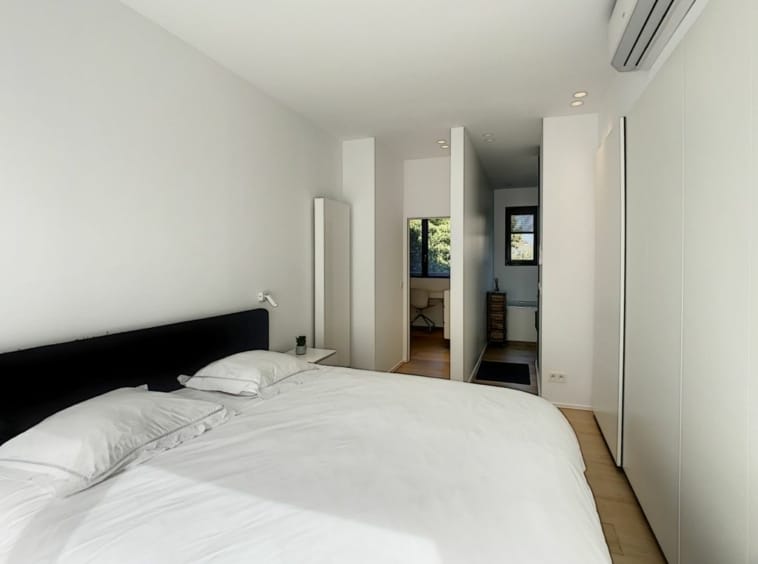 Chambre minimaliste moderne avec literie blanche, lumière naturelle et vue sur la nature extérieure.