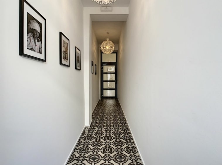 Couloir lumineux avec carrelage patterné, galerie photo, porte vitrée noire et chandelier moderne.