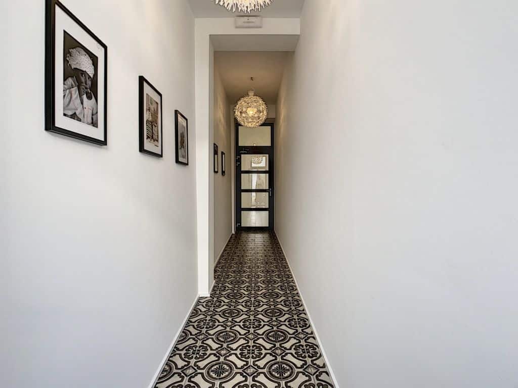 Couloir lumineux avec carrelage patterné, galerie photo, porte vitrée noire et chandelier moderne.