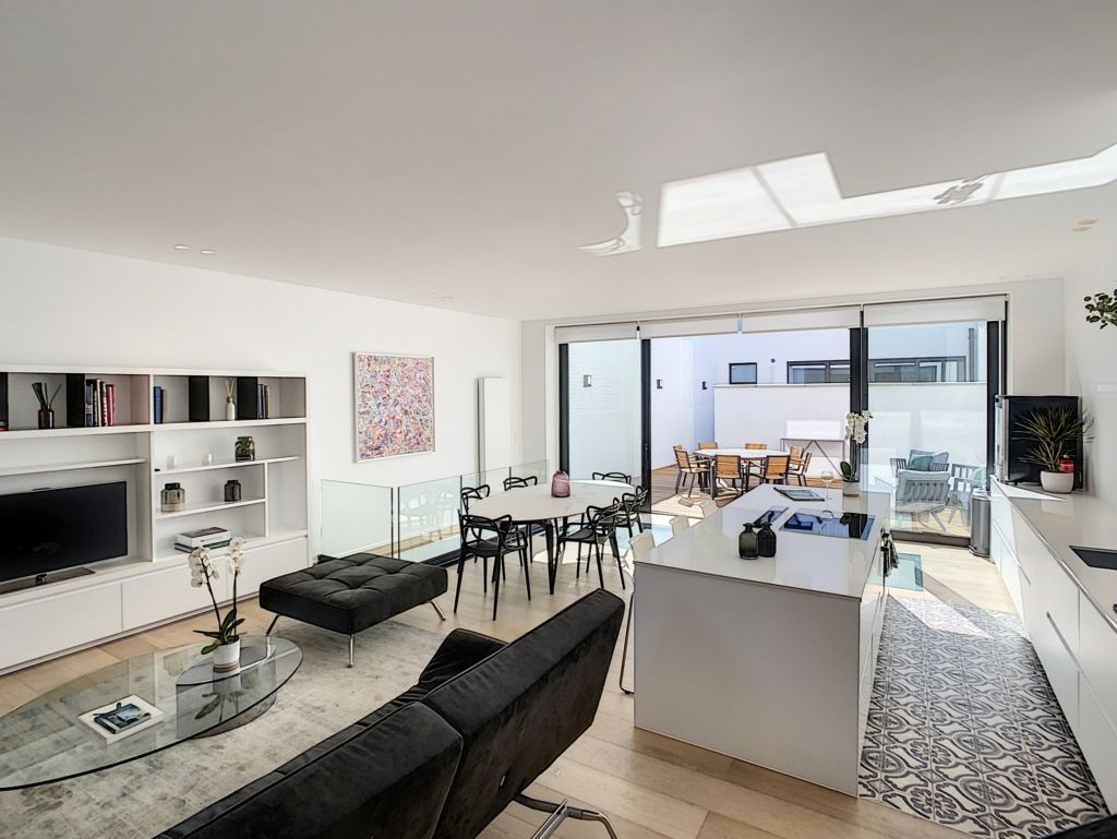 Salon moderne lumineux avec cuisine ouverte, canapé noir, terrasse ensoleillée, espace convivial et élégant.