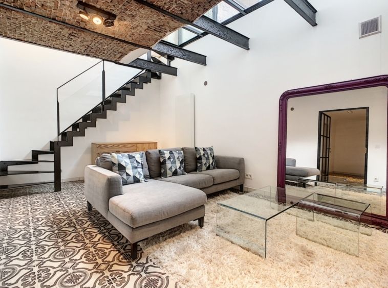 Salon moderne lumineux avec canapé gris, grand miroir violet, escalier minimaliste et décoration élégante.