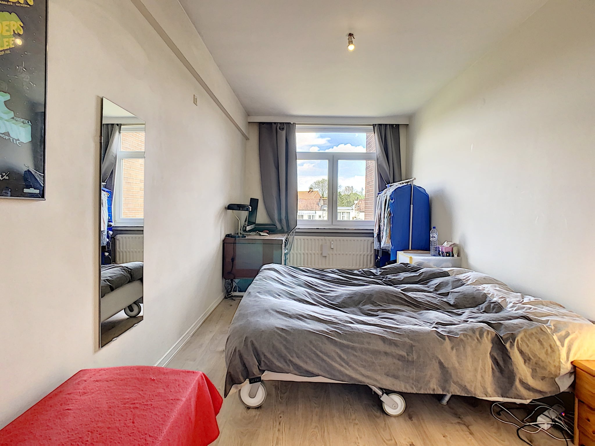 Chambre minimaliste lumineuse avec lit, bureau, armoire bleue, banc rouge et grande fenêtre naturelle.
