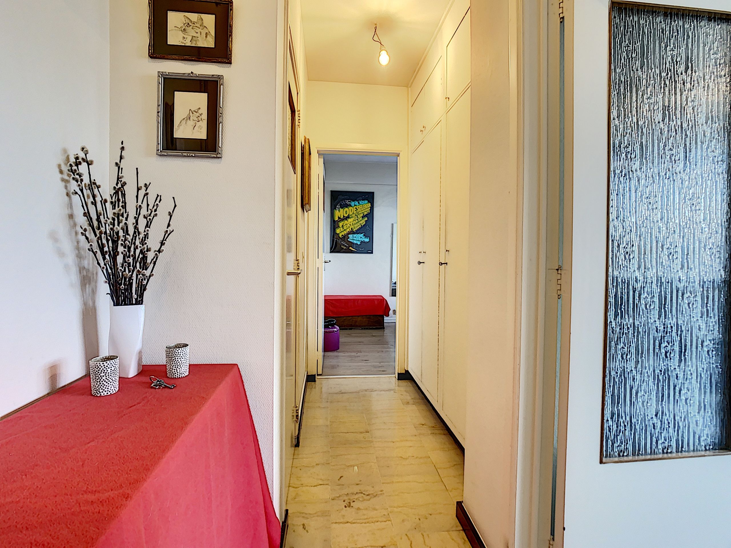 Couloir lumineux avec décorations, porte blanches, table rouge et chambre colorée au fond.
