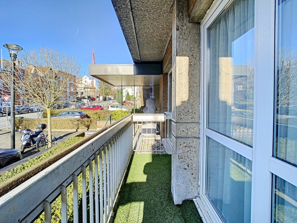 Balcon moderne avec gazon artificiel, vue sur rue animée, grandes fenêtres et ambiance urbaine.