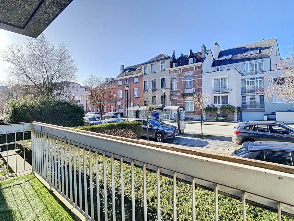 Balcon urbain calme avec vue sur rue bordée d’arbres nus, bâtiment en brique rouge, ciel bleu.