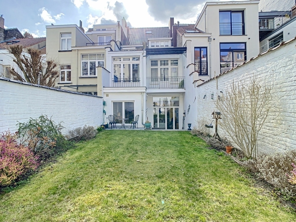 Maison moderne avec jardin verdoyant, grandes fenêtres et balcon, dans un cadre urbain paisible.