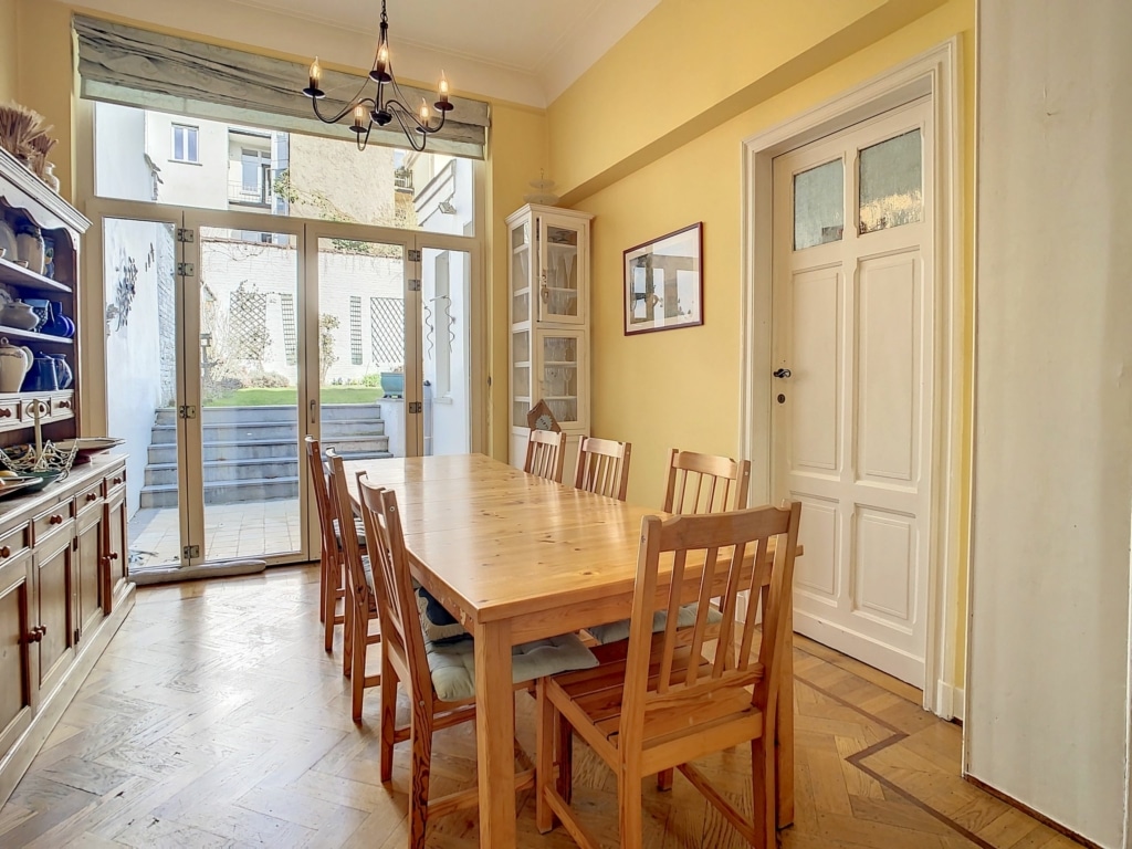 Salle à manger lumineuse avec table en bois, murs jaunes, décor bleu et vue terrasse naturelle.