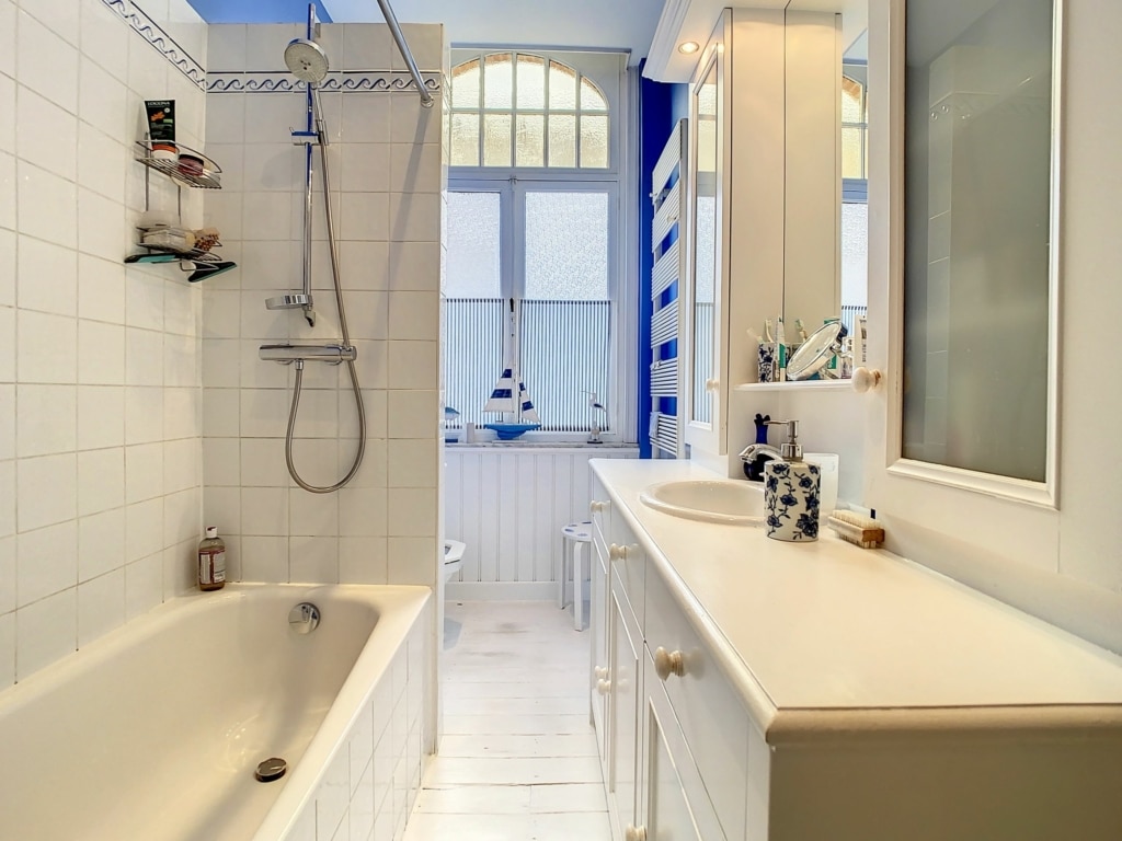 Salle de bain moderne lumineux, avec baignoire, douche, mobilier blanc épuré et large fenêtre naturelle.