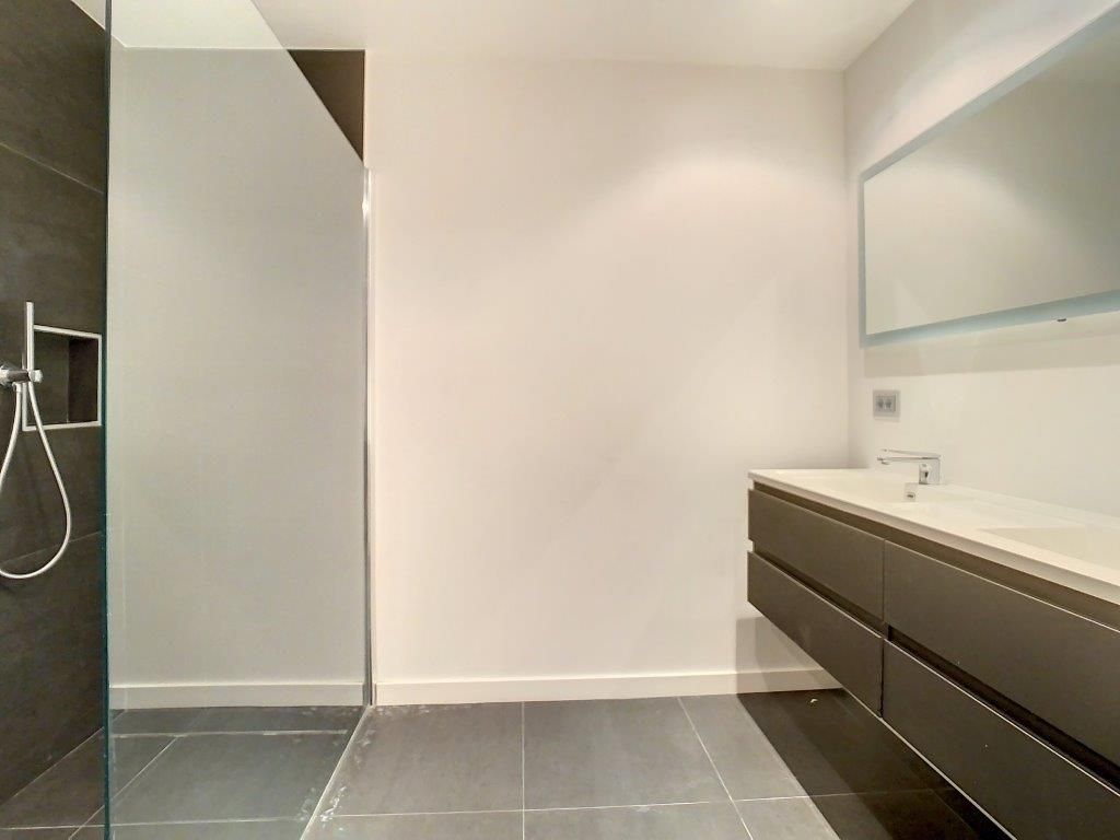 Salle de bain moderne minimaliste avec douche en verre, meuble en bois foncé, grand miroir et sol sombre.