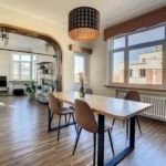 Salle à manger moderne avec table en bois, éclairage design, lumière naturelle et décoration végétale élégante.
