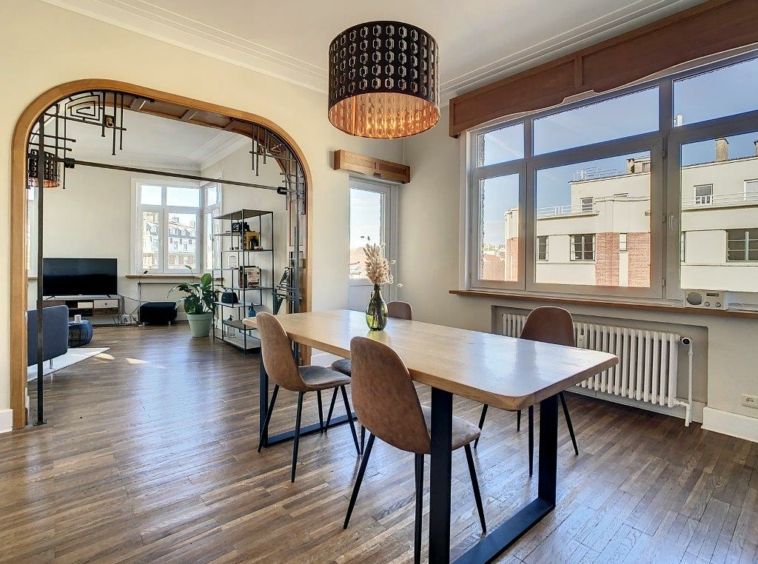 Salle à manger moderne avec table en bois, éclairage design, lumière naturelle et décoration végétale élégante.