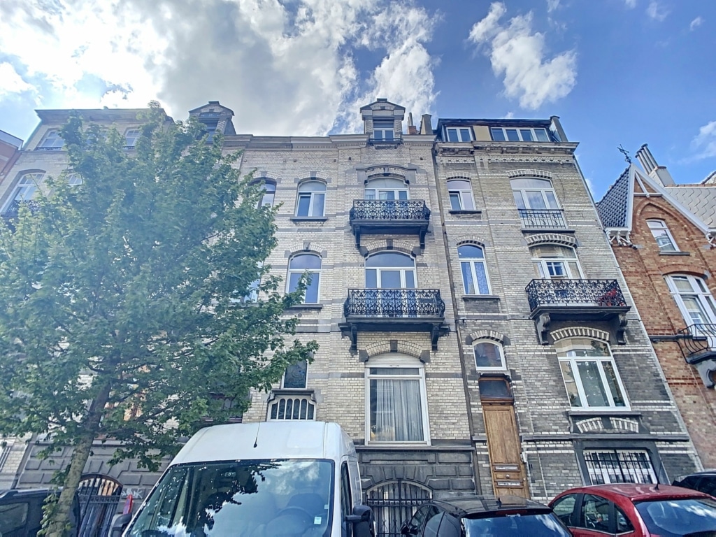 Façade ancienne en pierre avec balcons en fer forgé, fenêtres blanches, arbre et ambiance urbaine.