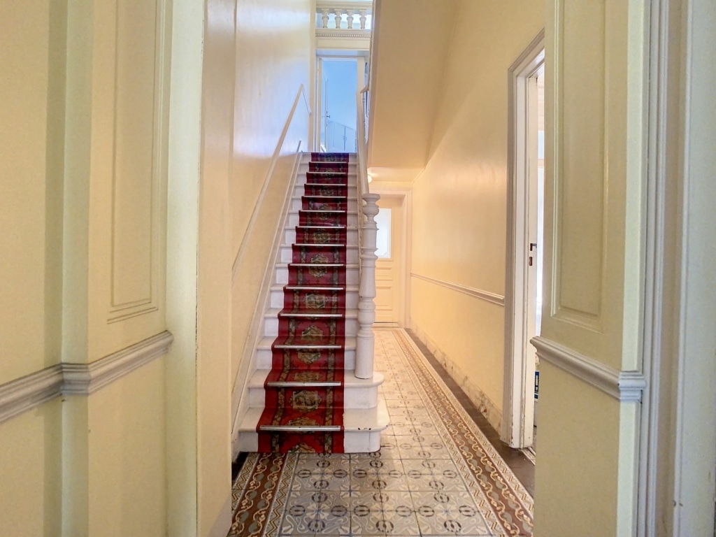 Couloir chaleureux avec escalier classique, tapis rouge et lumière naturelle, décoration élégante.