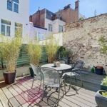 Terrasse urbaine ensoleillée avec mobilier moderne, plantes vertes et mur en briques.
