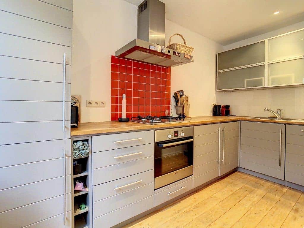 Cuisine moderne avec faïence rouge, meubles gris, plan en bois chaud et sol clair.