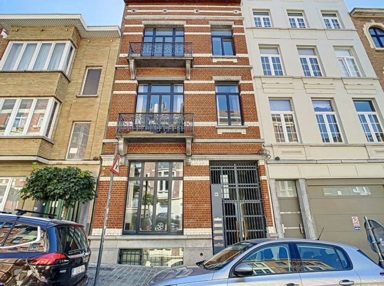 Immeuble en briques colorées, balcon en fer forgé, rue pavée animée avec voitures et verdure.