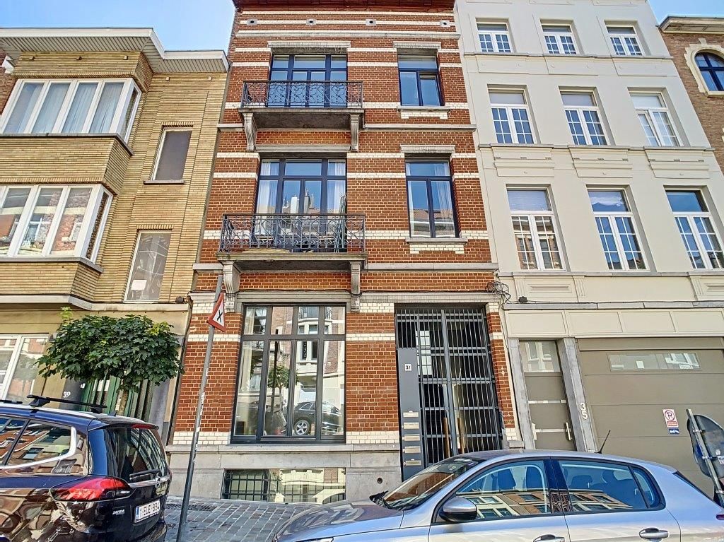 Immeuble en briques colorées, balcon en fer forgé, rue pavée animée avec voitures et verdure.
