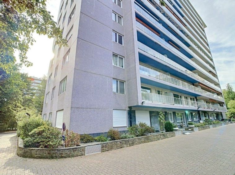 Résidence moderne avec balcons en verre, façade gris clair, jardins soignés et vue nature.