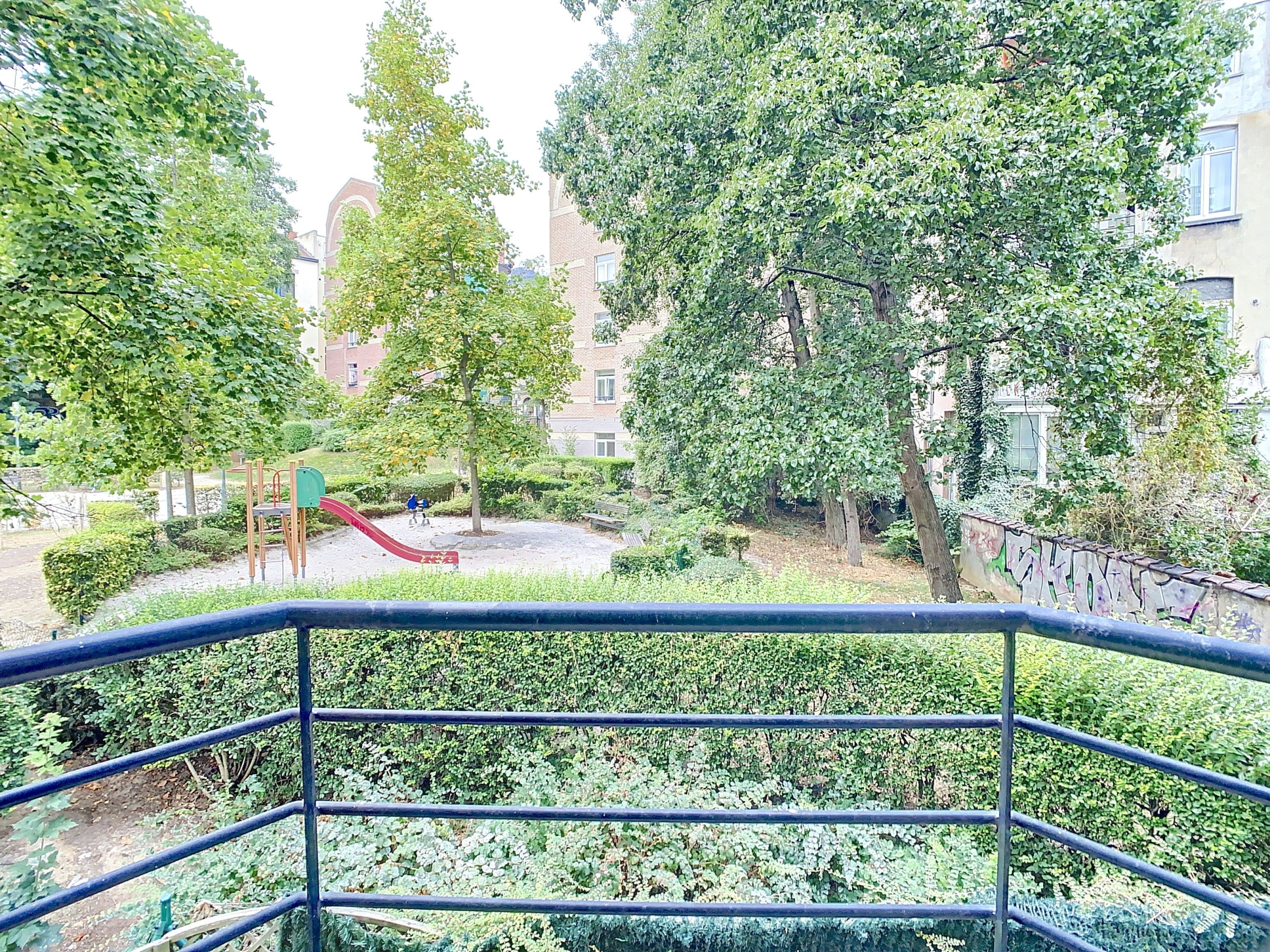 Balcon offrant vue sur jardin verdoyant et aire de jeux urbaine avec toboggan rouge.
