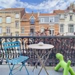 Balcon urbain coloré avec mobilier, vue pastel, ciel bleu, plantes, ambiance détendue et conviviale.
