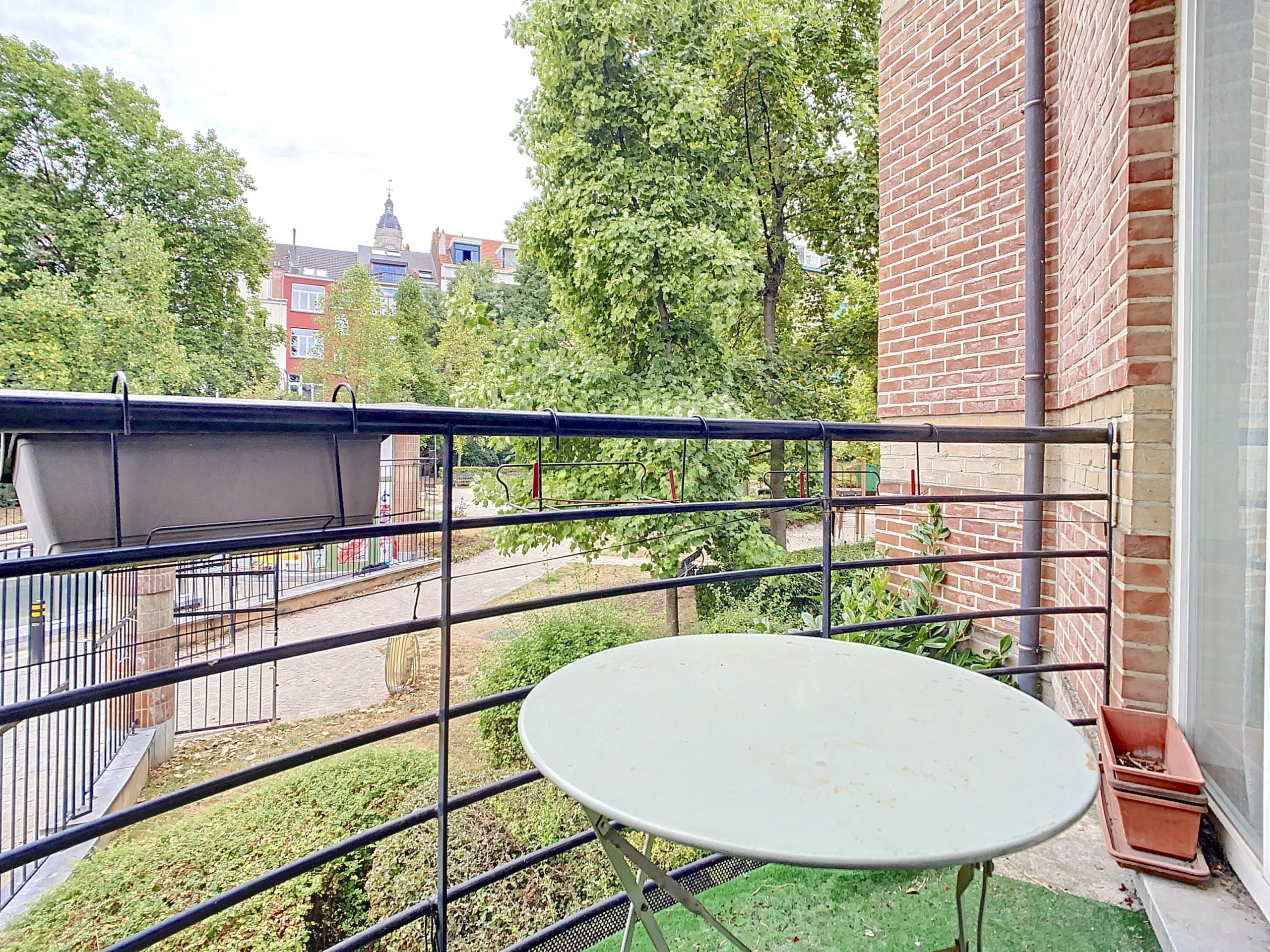 Balcon paisible avec table ronde, verdure, plantes en pot et vue sur jardin et quartiers voisins.