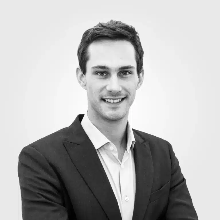 Homme professionnel en costume noir, sourire chaleureux et posture confiante, fond neutre.