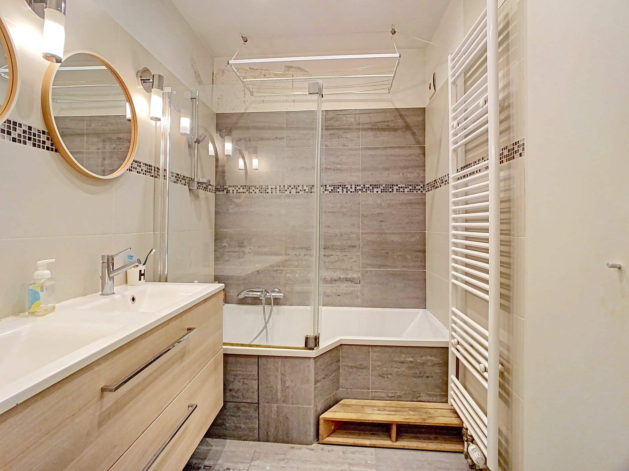 Salle de bain moderne en gris, bois, baignoire en verre, miroir rond, radiateur vertical blanc.