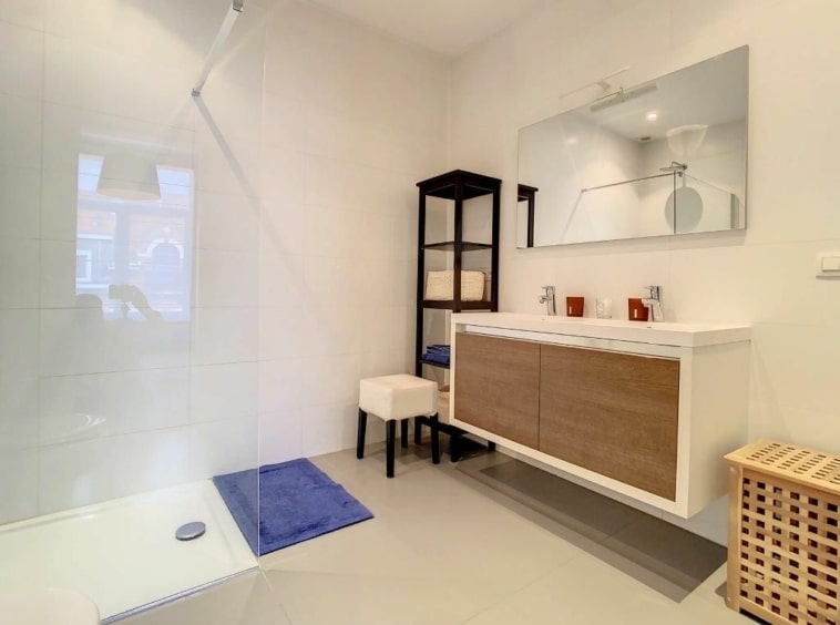 Salle de bain moderne lumineuse avec douche en verre, mobilier en bois clair et rangements élégants.