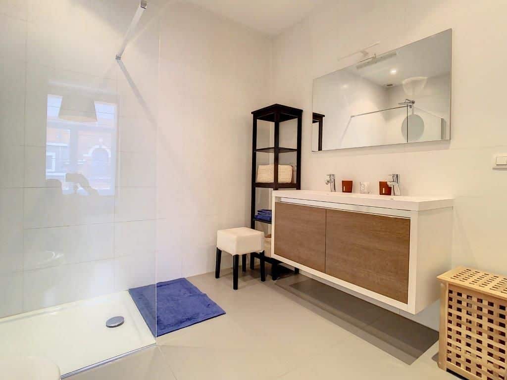 Salle de bain moderne lumineuse avec douche en verre, mobilier en bois clair et rangements élégants.