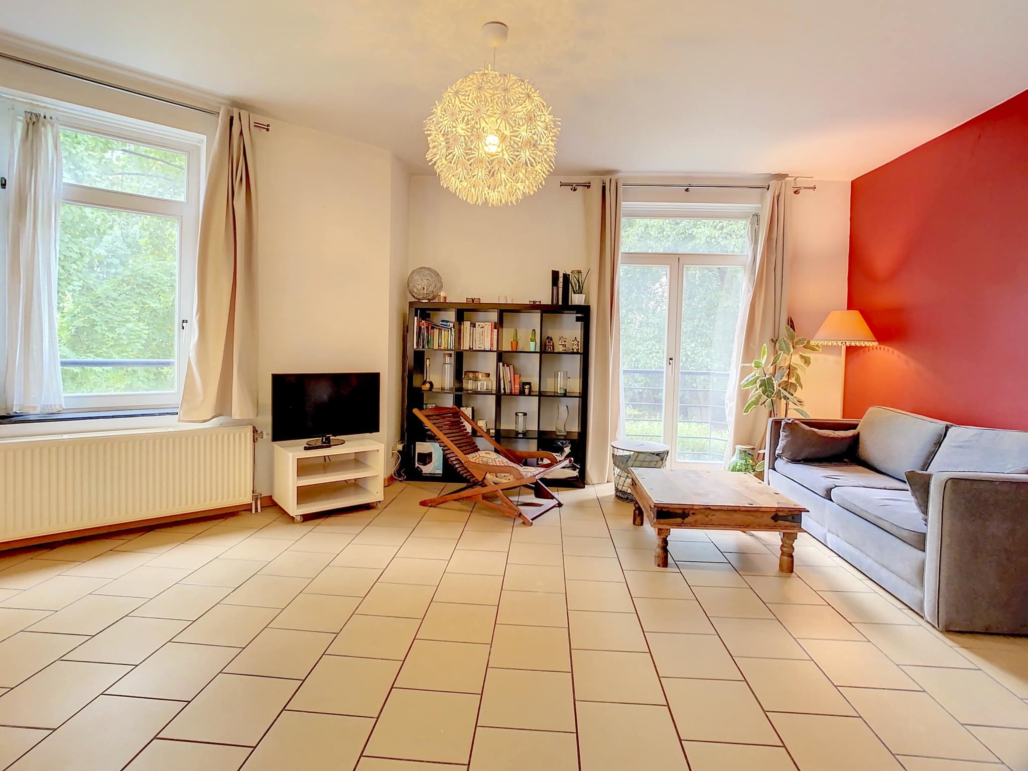 Salon moderne et chaleureux avec mur rouge, canapé gris, lumière naturelle et ambiance conviviale.