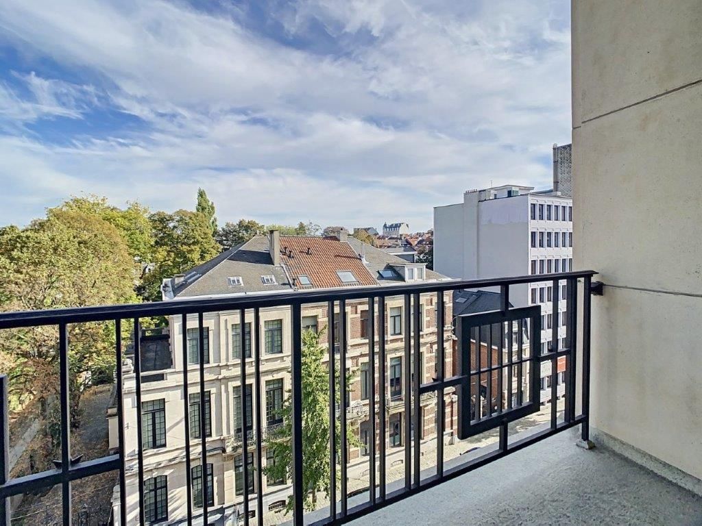 Vue de balcon urbain avec bâtiments historiques, modernes, arbres verts et ciel partiellement nuageux.
