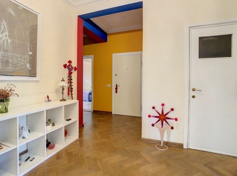 Couloir moderne avec parquet en chevron, murs colorés, étagères décoratives, porte vitrée et sculpture rouge.