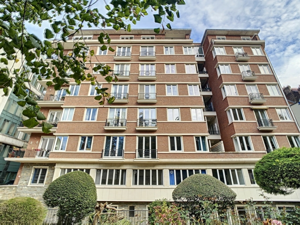 Immeuble résidentiel en brique rouge avec jardins, balcons en fer forgé et ambiance paisible.