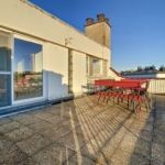 Terrasse urbaine ensoleillée avec mobilier rouge, vue dégagée, grande porte vitrée, ambiance contemporaine.