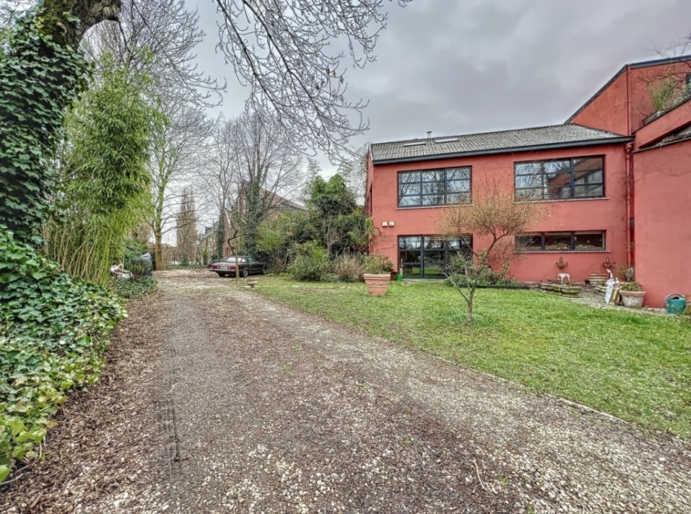 Maison contemporaine rouge dans un jardin paisible avec chemin en gravier et végétation luxuriante.