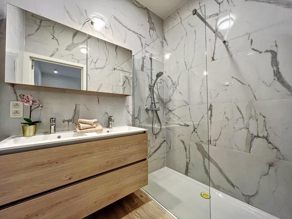 Salle de bain moderne en marbre blanc veiné gris avec meuble en bois et orchidée.