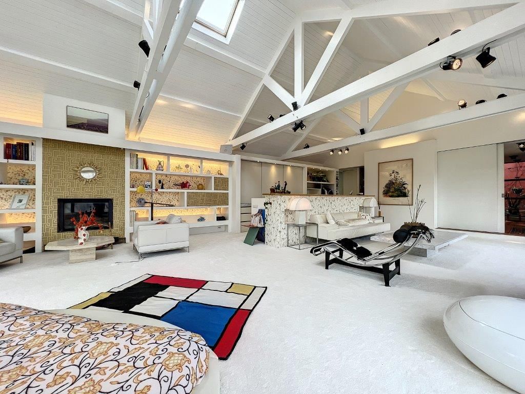 Salon moderne lumineux avec mobilier design, tapis coloré, mur décoratif et coin cuisine minimaliste.