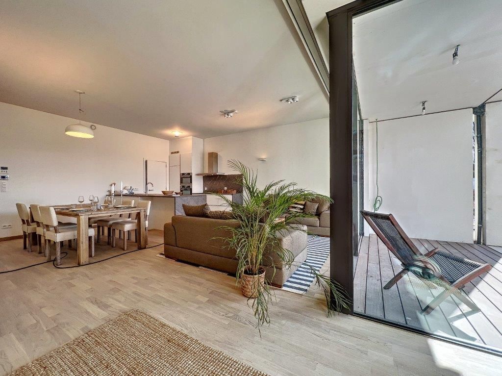 Salon moderne lumineux avec canapé, cuisine équipée, large fenêtres et espace extérieur confortable.