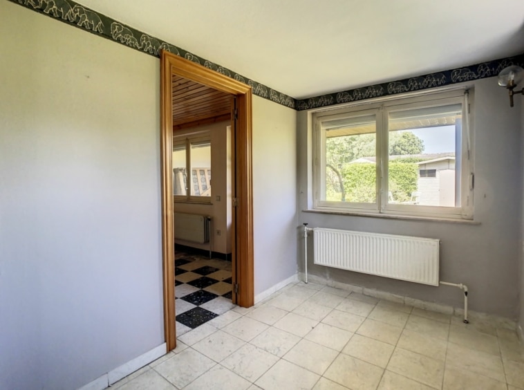 Pièce lumineuse avec murs gris, fenêtre, radiateur, carrelage clair et porte en bois visible.