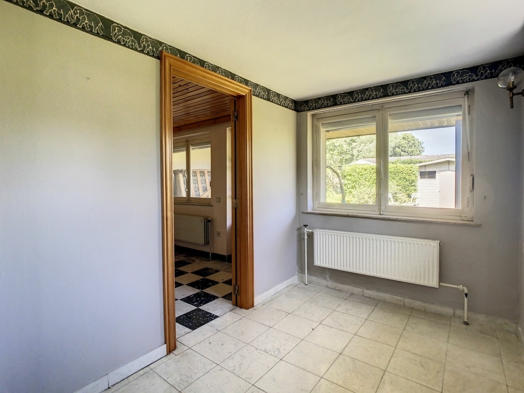 Pièce lumineuse avec murs gris, fenêtre, radiateur, carrelage clair et porte en bois visible.