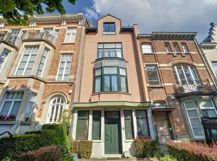 Maison urbaine moderne avec grand bay window, façade claire et terrasse sur toit, entourée de verdure.