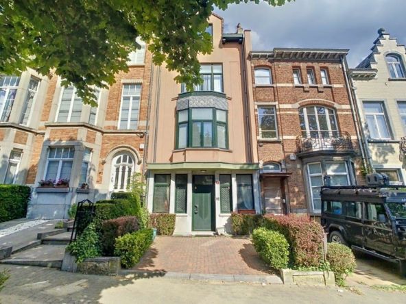 Rue urbaine avec bâtiments en briques, voitures vintage, jardins soignés et ciel nuageux.