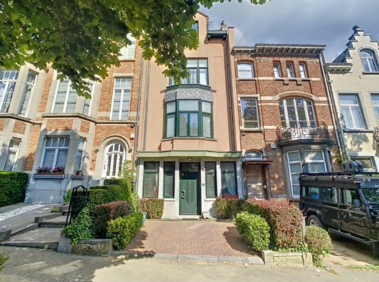 Rue urbaine avec bâtiments en briques, voitures vintage, jardins soignés et ciel nuageux.