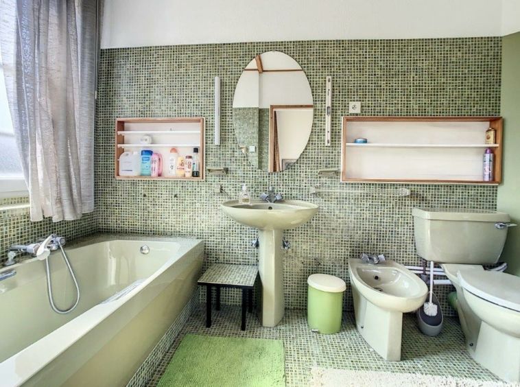 Salle de bain moderne avec tuiles vertes, baignoire, lavabo, miroir rond, étagères en bois.