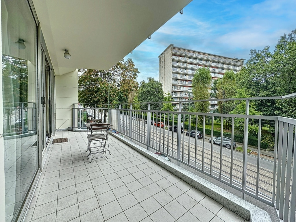Balcon lumineux avec vue sur jardin urbain et architecture moderne sous ciel partiellement nuageux.