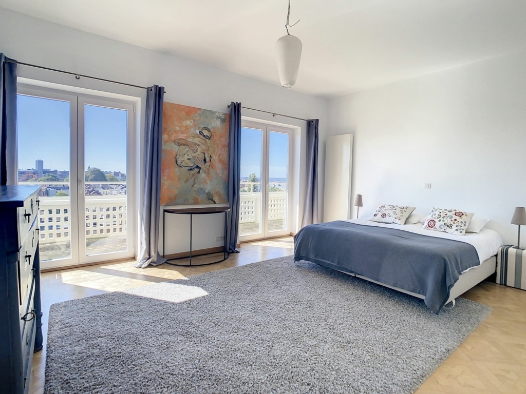 Chambre lumineuse et moderne avec lit gris, fenêtres naturelles, art coloré et atmosphère apaisante.