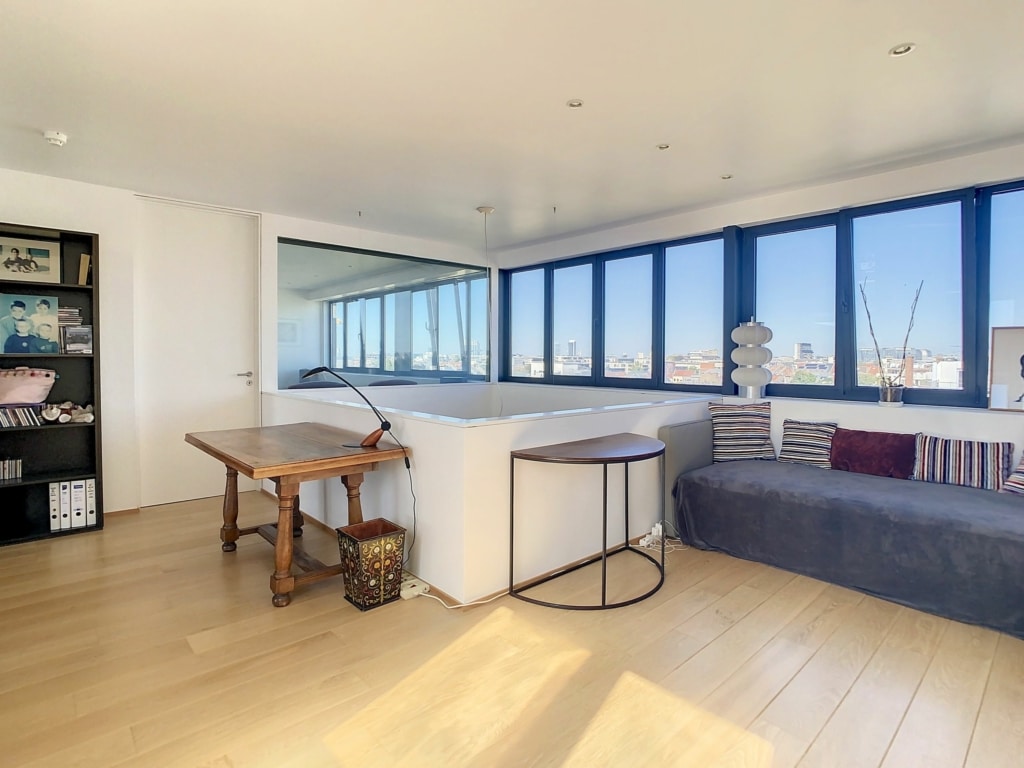 Salon lumineux avec vue urbaine, mobilier moderne, canapé gris, étagère noire et ambiance chaleureuse.