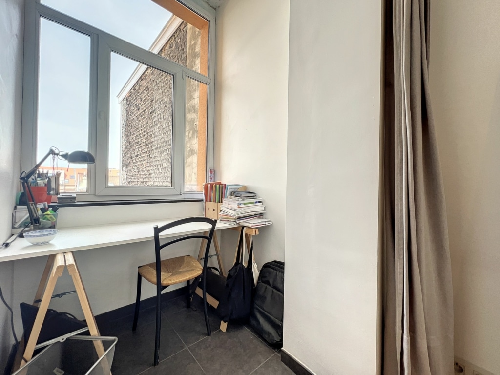 Coin de bureau lumineux minimaliste avec vue, bureau en bois clair, chaise noire, sacs, et rideau léger.
