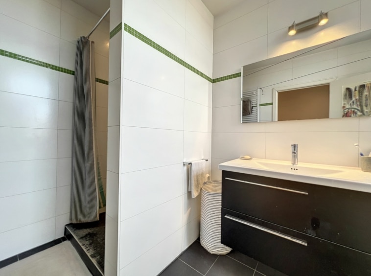 Salle de bain moderne avec carrelage blanc, douche en verre, meuble en bois foncé et miroir large.