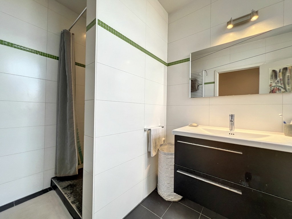 Salle de bain moderne avec carrelage blanc, douche en verre, meuble en bois foncé et miroir large.